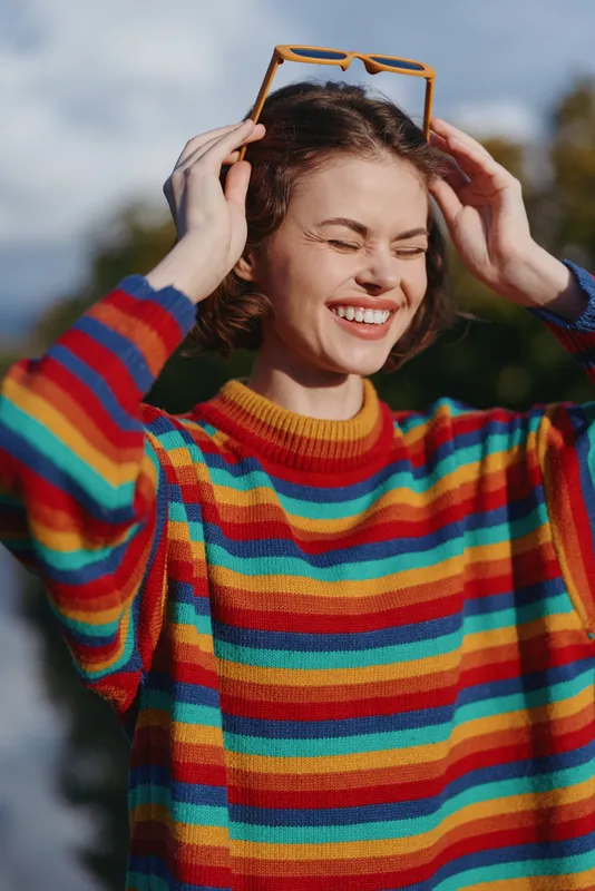 woman smiling in a rainbow sweater outdoors, young woman adjusting sunglasses on head with eyes closed, casual outfit and joyful expression in natural light lifestyle portrait.