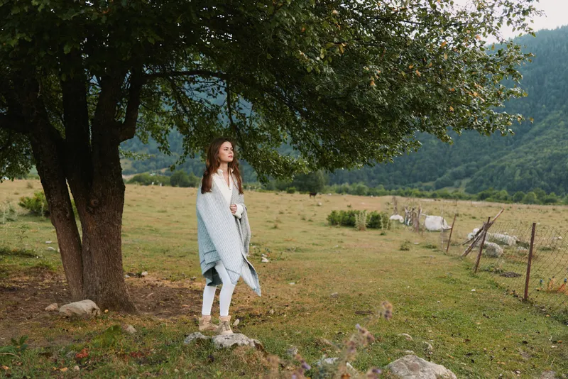 Young woman wrapped in blanket stands under tree in countryside field near wooden fence with cows grazing in background on cloudy day, enjoying peaceful lifestyle.