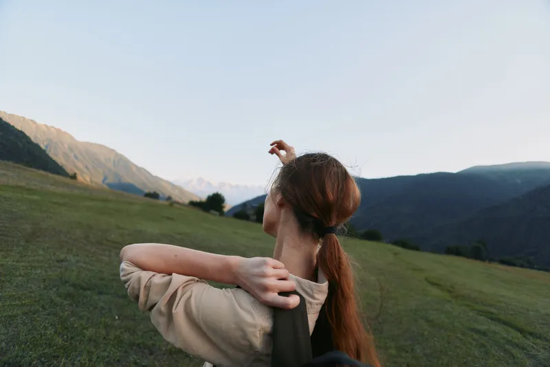 Woman in a grassy hillside landscape enjoying outdoor lifestyle, relaxed pose, looking toward distant mountains, casual attire, golden hour light, sense of freedom and exploration