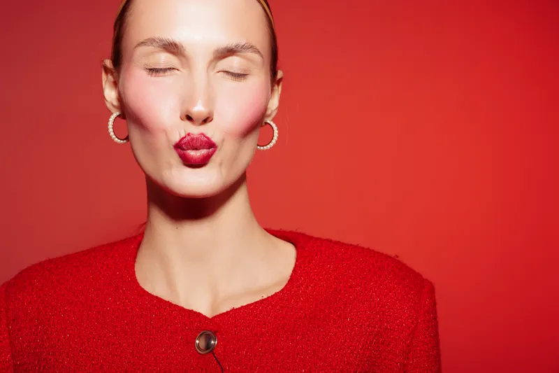 Young woman in red sweater with closed eyes blows a kiss on red background. Concept of affection, beauty, and playful emotions in studio portrait photography.