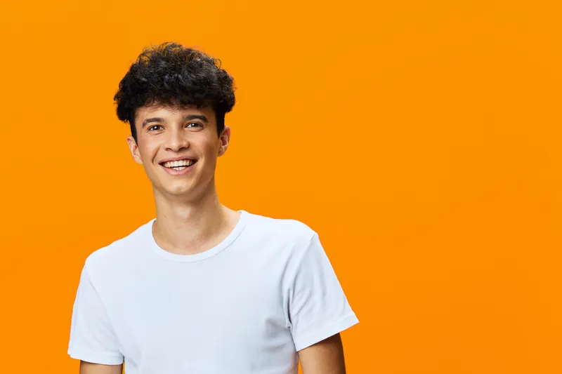 smiling man in white t-shirt posing against vibrant orange background with curly hair and youthful happy expression, casual style portrait in bright studio lighting