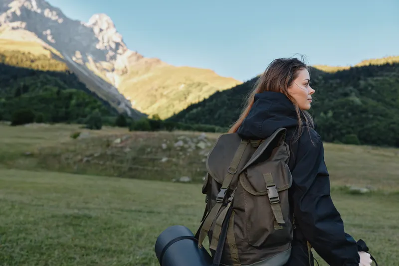 A traveler with a backpack wanders across a grassy meadow beneath towering mountains, wearing a dark jacket. The scene conveys solitude, exploration, and calm energy, inviting a connection with