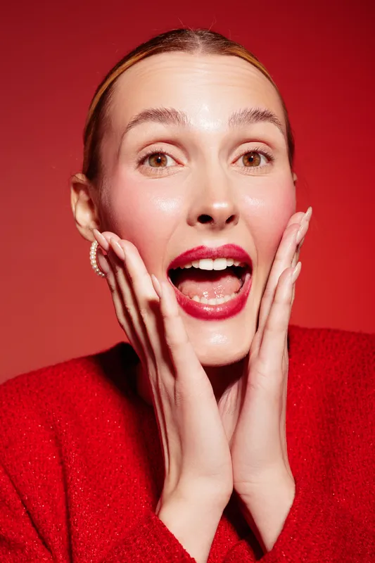Surprised woman with red lips and shiny skin touches her face with both hands on red background. Concept of beauty, skincare, emotion, and natural makeup in studio lighting.