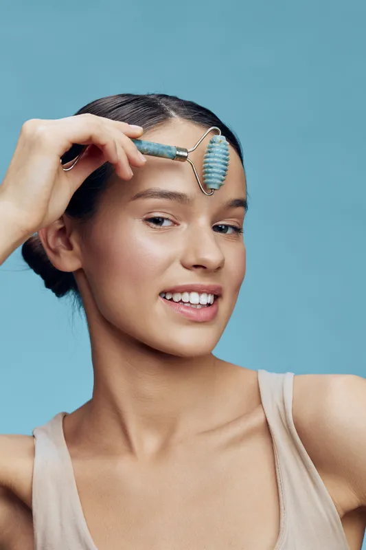 Smiling young woman using a facial roller for skincare on a light blue background, promoting healthy beauty and self care routines