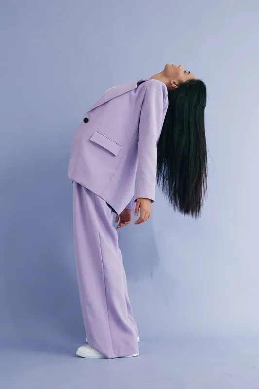 Young woman in lilac suit leans backward with long black hair, standing against pastel purple background portraying grace and modern fashion style.