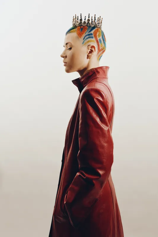 woman with colorful shaved hair and crown wearing red leather coat standing sideways on neutral background, stylish and bold alternative fashion look