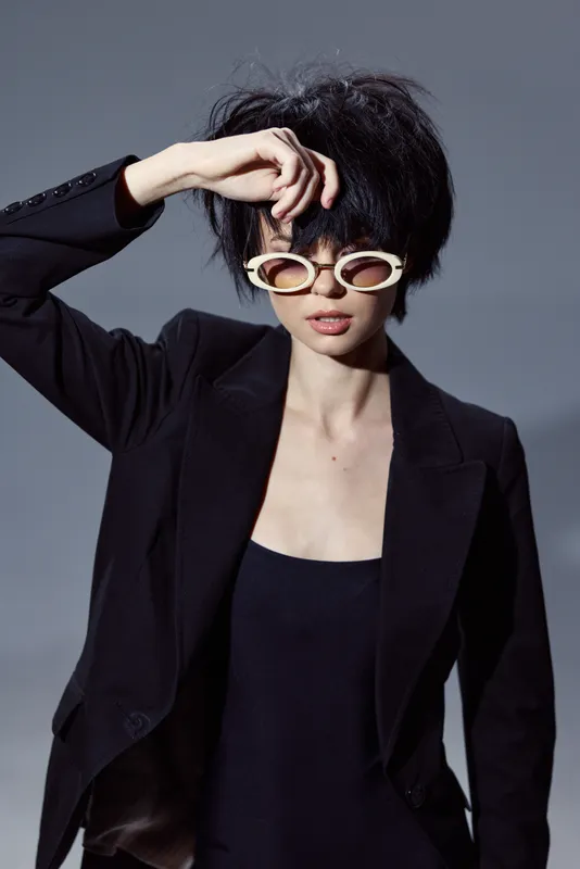 Stylish woman with short hair wearing black blazer and unique sunglasses posing in studio.
