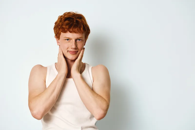 Young man with red hair and fair skin wearing white tank top posing with hands on face against white background. Portrait of confident male model with freckles.