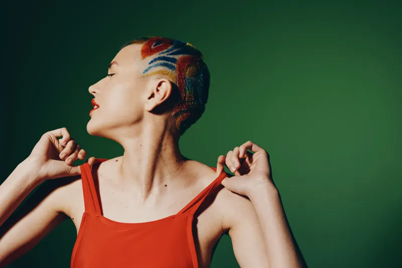 woman with colorful hair and vibrant patterned shaved head wearing red sleeveless top posing against green background, stylish and artistic portrait