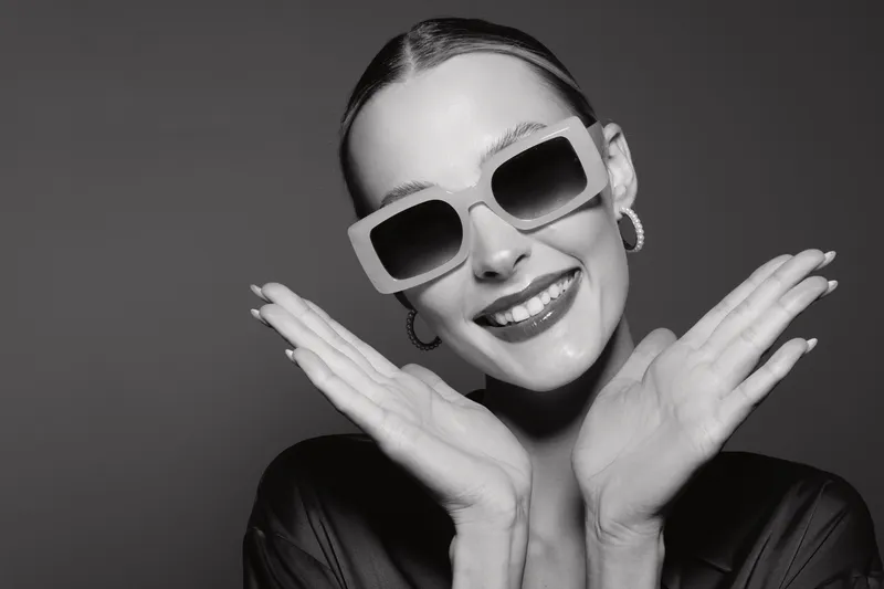Smiling young Caucasian woman with light skin wearing large sunglasses and earrings poses happily with hands near face on dark background.