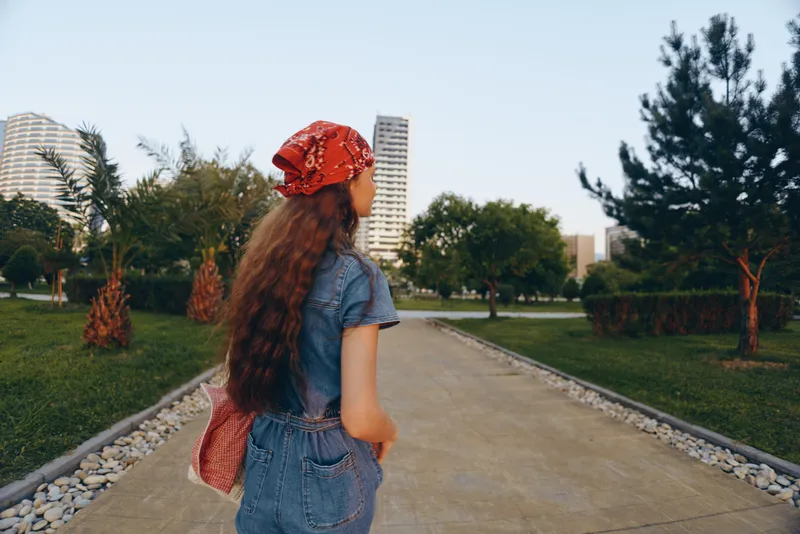 lifestyle woman in modern boho-western denim street style wearing red bandana walking in urban park with green trees and buildings, film color aesthetic