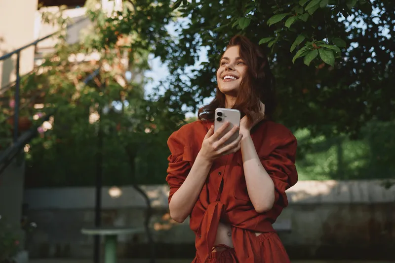 Young woman in a red dress smiles while holding a smartphone outdoors, enjoying a bright afternoon in a leafy urban park, capturing a casual lifestyle moment