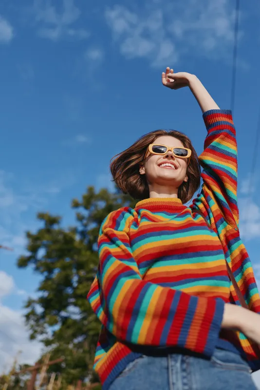 Woman in rainbow sweater and sunglasses smiling outdoors under blue sky, casual joyful young adult posing with raised arm in park setting for lifestyle, travel and fashion content.
