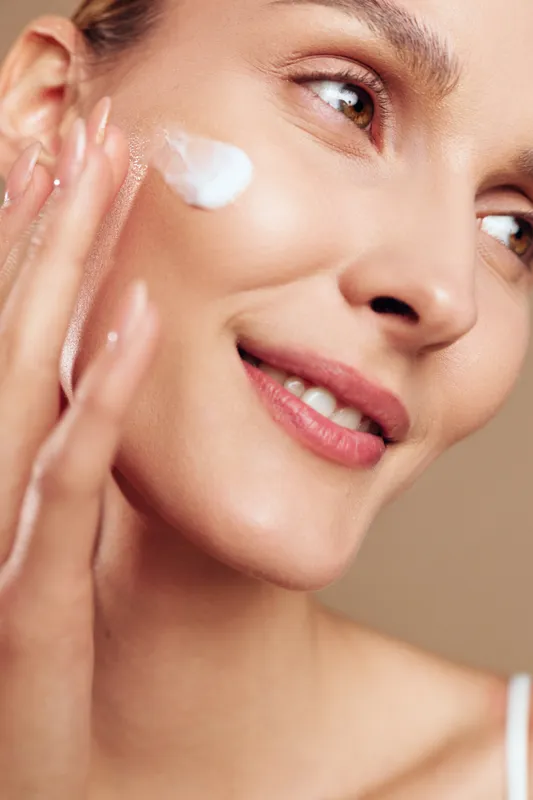 Close-up of smiling woman applying white face cream on cheek with fingers. Natural skincare routine concept with soft lighting and warm beige background. Healthy skin care beauty.