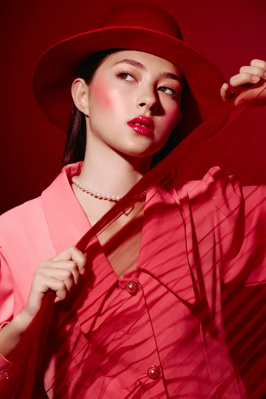 Young woman in bright red hat and pink outfit poses creatively while holding red fabric. Studio portrait with striking makeup in a bold fashion and beauty concept.