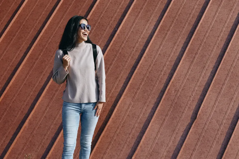 Outdoor fashion scene with a smiling woman wearing sunglasses, a light sweater and jeans, standing beside a rust diagonal wall, showcasing casual style, confident pose, and a sunlit urban backdrop.
