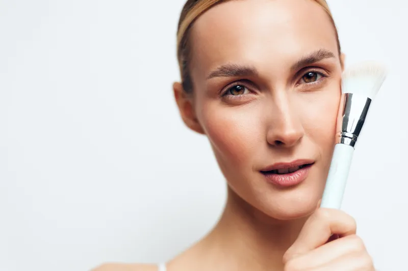 Closeup portrait of woman with natural skin holding makeup brush near face applying powder with soft brush on smooth complexion isolated on white background.