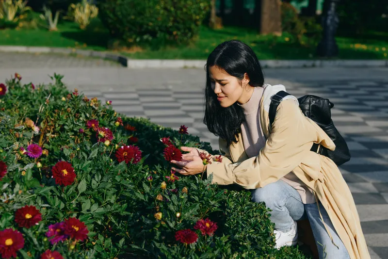 Woman kneeling beside vibrant flowers in a garden, admiring blossoms during a sunny outdoor stroll, wearing a light coat and backpack in casual style under clear sky
