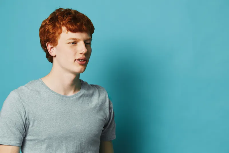 Young man with red hair and casual gray t shirt standing against blue background, looking to side with neutral expression in studio portrait.