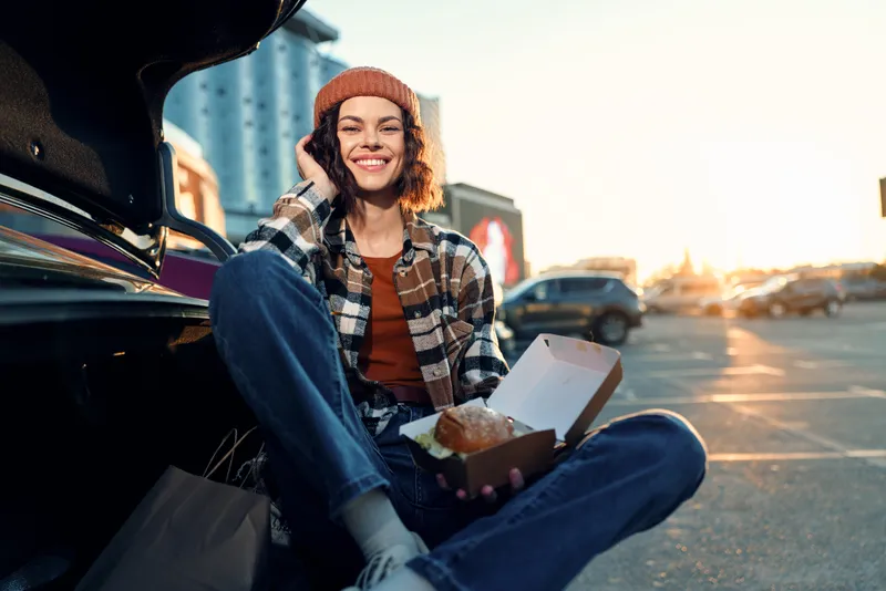 Woman sitting by car trunk in parking lot, holding food box and smiling at golden hour glow; candid lifestyle portrait with authenticity, mindful living and emotional storytelling vibe.