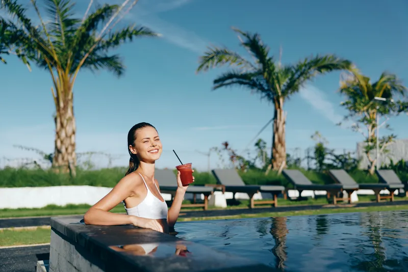 A smiling young woman in a white swimsuit relaxes in an outdoor pool holding a red drink with a straw. Palm trees and lounge chairs are visible under a clear blue sky in the background.
