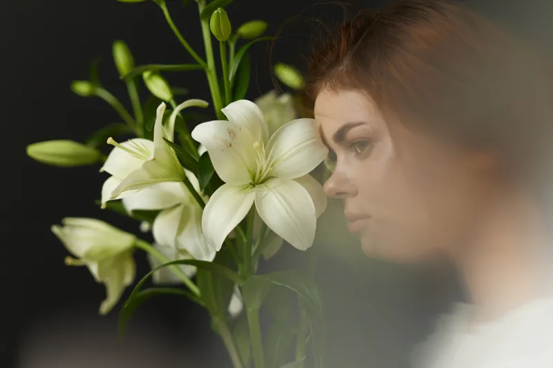Young woman with brown hair interacting with white lilies, showcasing emotions in a serene environment with a soft focus background
