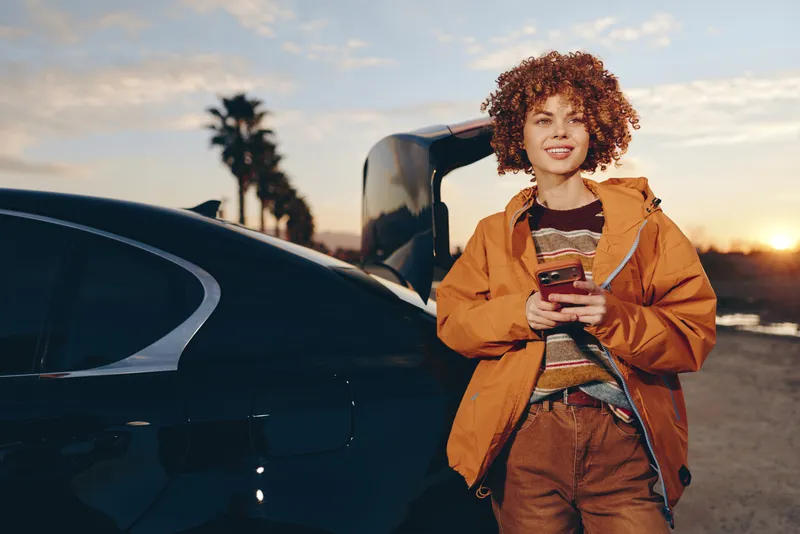 Woman outdoors near car at sunset, holding phone in hands, curly hair and confident smile, warm golden light, casual jacket, relaxed posture, urban street with palms, lifestyle travel moment