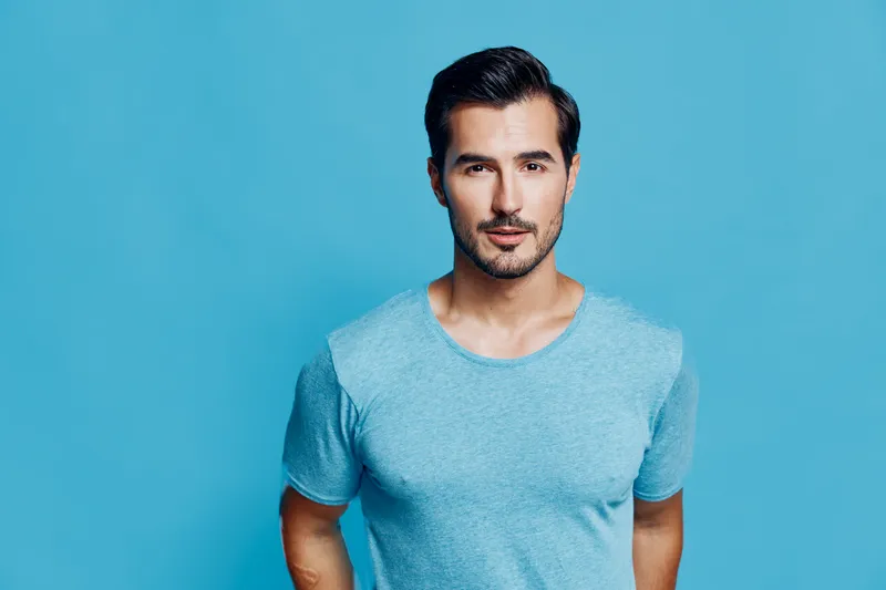 Confident man wearing a light blue t-shirt poses against a solid turquoise background, showcasing an engaging expression and stylish appearance. Portrait concept