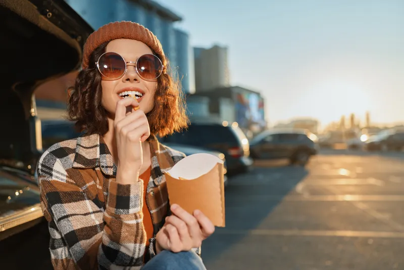 woman portrait popcorn car sunset sunglasses beanie parking Candid authenticity and lifestyle scene of a smiling young woman eating popcorn by an open trunk in golden hour glow, mindful living and