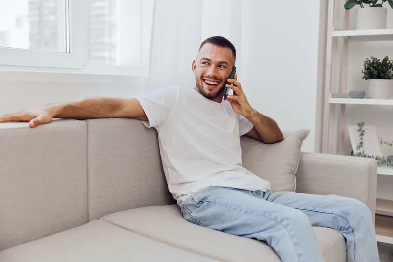 Smiling man talking on the phone, relaxed atmosphere, modern living room, casual attire, bright colors, lifestyle concept, enjoying conversation and comfort.