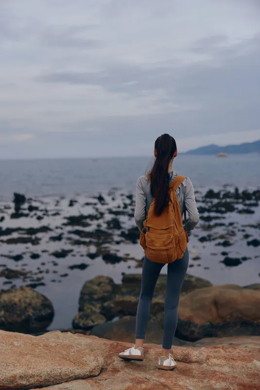 Young woman with a backpack standing on rocky shoreline, gazing at the serene sea, embodying a sense of adventure and tranquility in nature