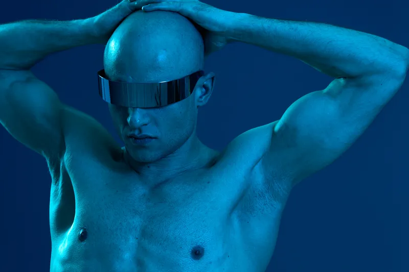 Stylish man with futuristic glasses poses confidently, showcasing a strong physique. The vibrant blue lighting adds a bold atmosphere to the striking composition.
