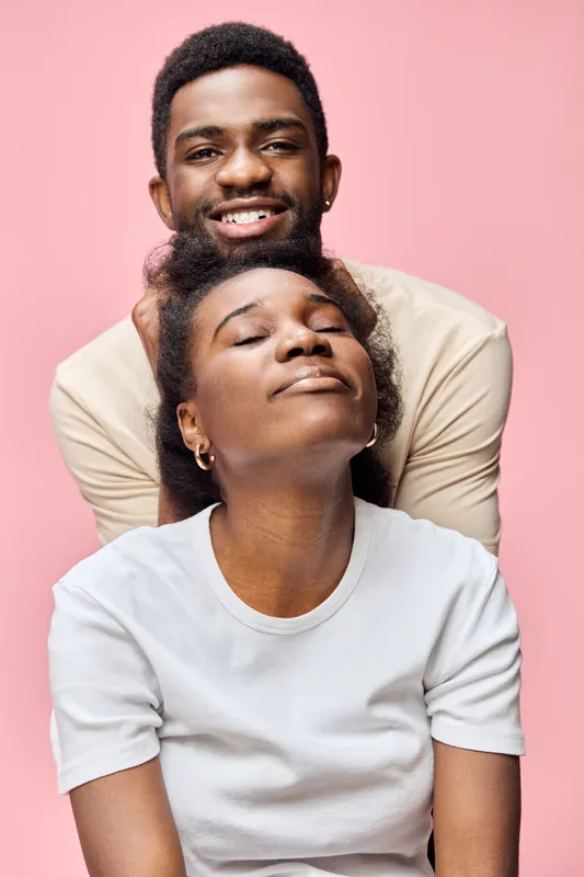 A joyful couple showcasing love and happiness against a soft pink background, featuring a relaxed woman in a white shirt and a smiling man behind her
