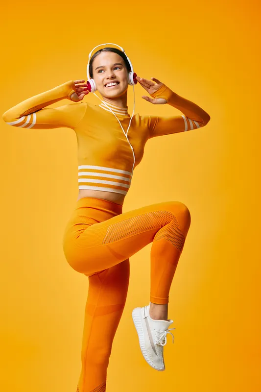 Energetic woman in bright athletic wear dancing with headphones against a vibrant yellow backdrop, capturing dynamic fitness movement, personal motivation, and an upbeat workout atmosphere full of