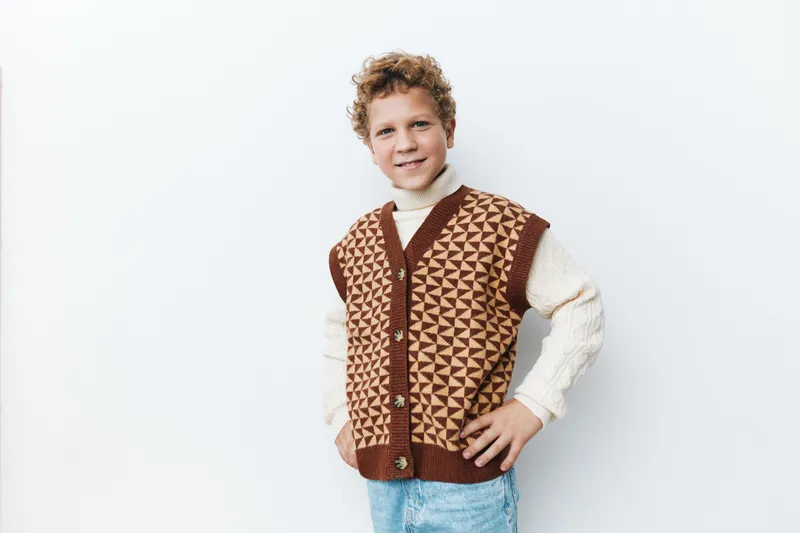 Young boy models a cozy knitted vest over a cream sweater, standing in a studio against a white backdrop, fashion forward, playful, confident, and warmly styled.