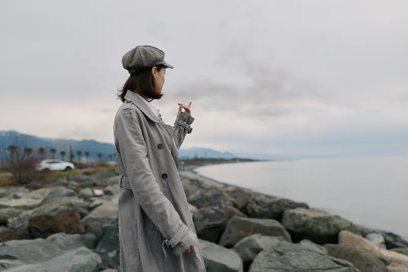 Young woman in checkered coat and cap smokes cigarette standing on rocky shore with ocean and cloudy sky in background. Concept of solitude, contemplation, and urban fashion outdoors.