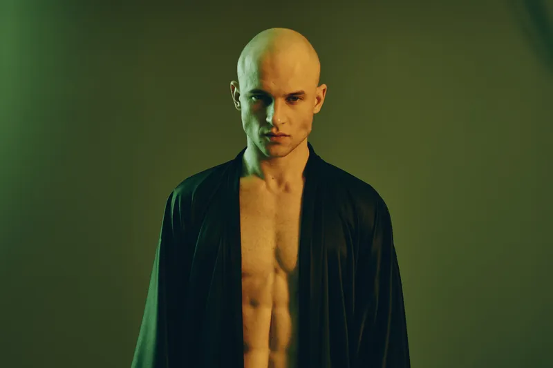 Portrait of a man with a strong posture. He has a shaved head and is dressed in a black robe against a light background with a serene mood.