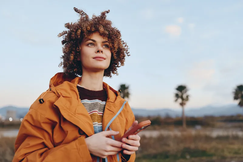 Woman outdoors, curly hair, orange jacket, smiling while using a smartphone, relaxed lifestyle moment, soft sunset light, open field and distant palms, natural colors, calm mood