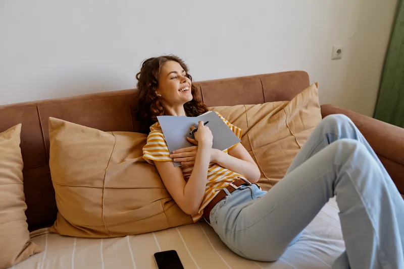 Smiling woman lifestyle home relaxation on a sofa, holding a notebook in a casual moment. Cozy living room scene with warm light, showcasing comfort, creativity, and everyday happiness.