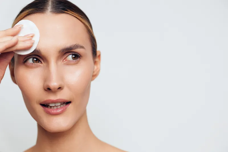 Young woman with brunette hair gently cleanses her face using a cotton pad against a plain light background, showcasing a skincare routine with a calm and focused expression.