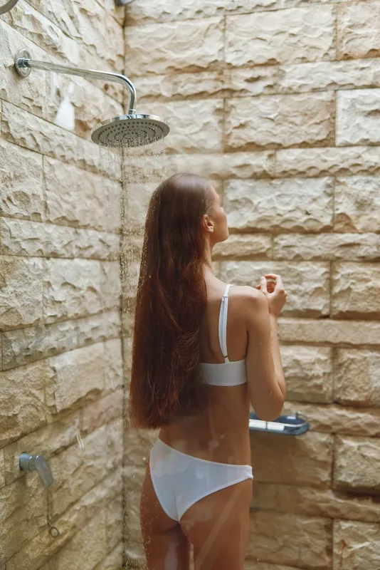 Woman showering outdoors in white underwear with natural stone wall background, long brown hair falling down her back, warm lighting, and relaxed mood.