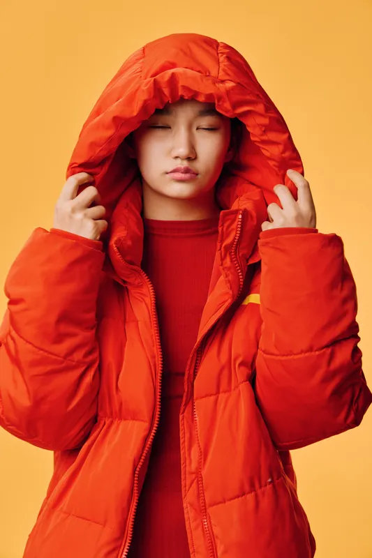 Young woman wearing red puffer jacket with hood on head, eyes closed, standing against orange background. Portrait of casual fashion, winter outerwear, cold weather and style concept.