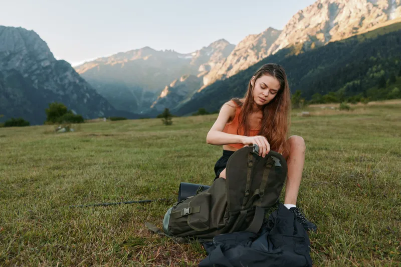 A young woman sits on a grassy field in a mountain landscape, carefully packing a rugged backpack. She wears casual clothes and looks calm, focused, and ready for outdoor exploration.