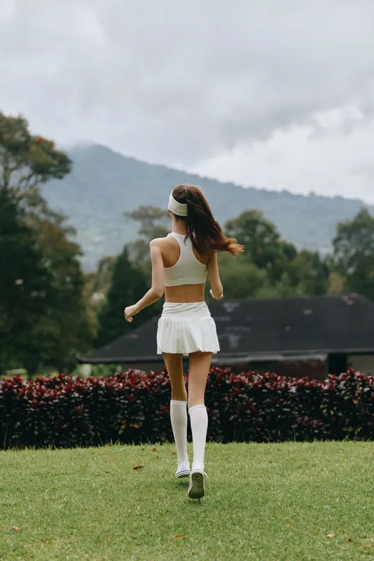 Young athletic female model in white sportswear jogging outdoors in a lush green landscape with mountains, embodying a healthy lifestyle and fitness enthusiasm