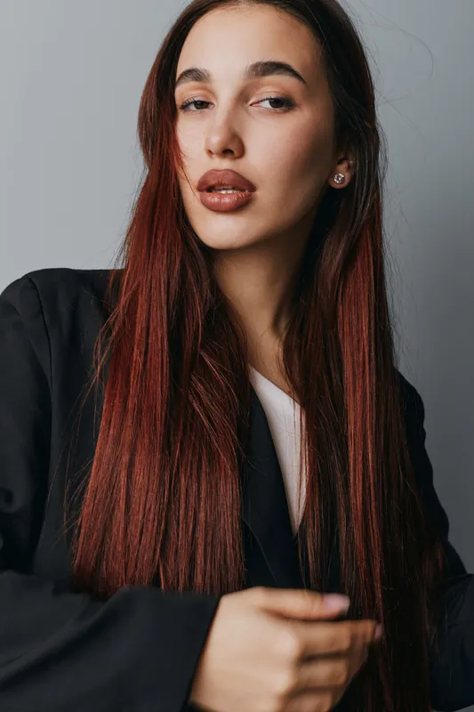 Young woman with long straight red hair wearing black blazer and white top posing with confident expression against gray background in studio portrait.