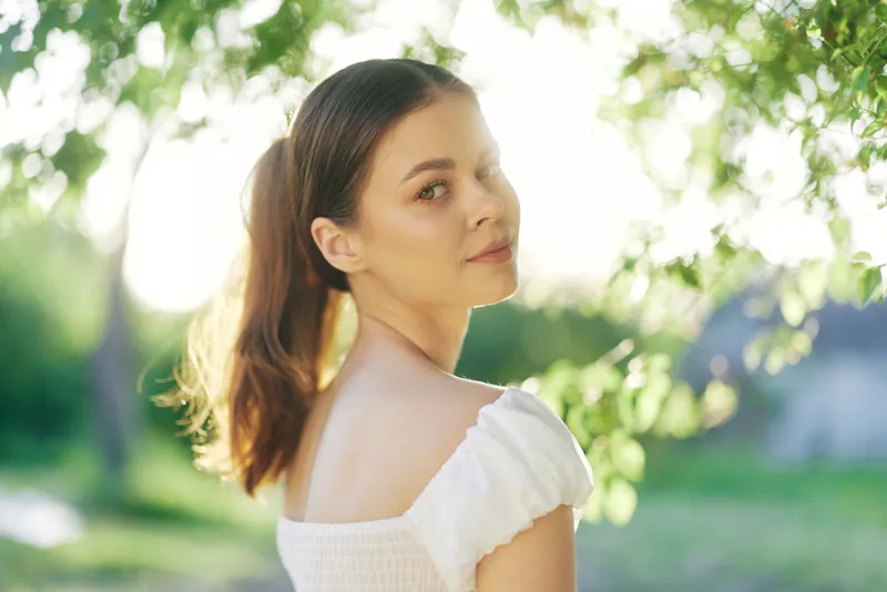 Young woman in white dress smiling softly among green leaves, capturing natural beauty and tranquility in a sunny outdoor setting
