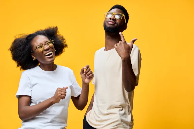 Happy young couple dancing on a vibrant yellow background, wearing stylish sunglasses, showcasing joy and positivity through their playful expressions
