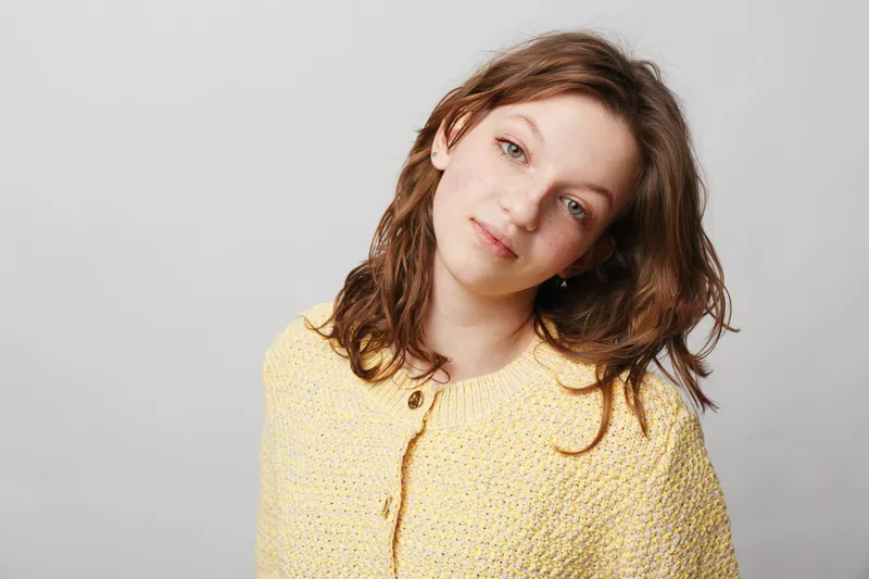 Young woman with brown hair wearing yellow knitted sweater looks gently at camera on grey background. Soft natural light highlights her calm and relaxed facial expression and casual style.