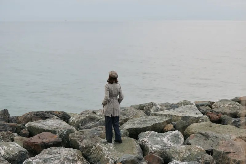 A woman in a coat and hat stands on rocky shore facing calm sea under overcast sky. The scene conveys solitude, contemplation, nature, and peaceful outdoor mood near water.