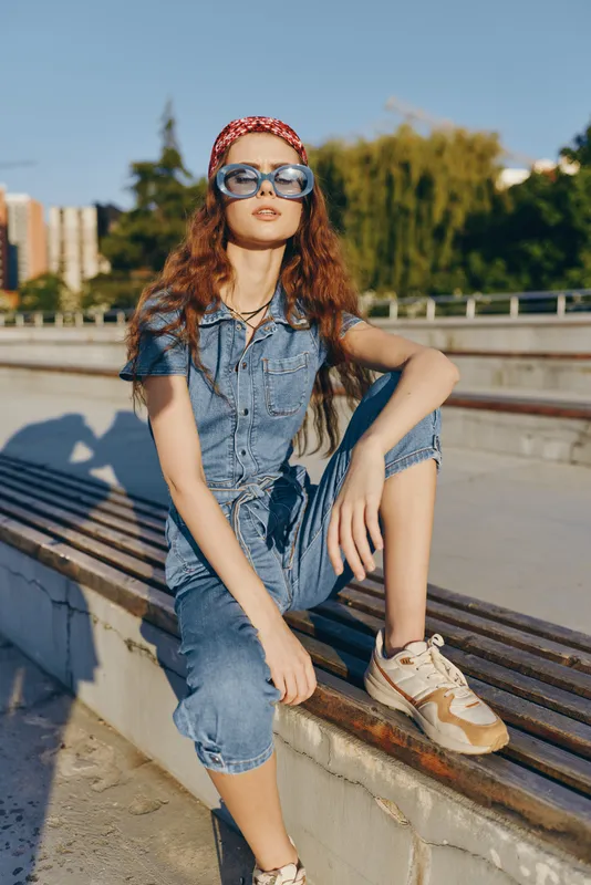 Lifestyle woman in modern boho-western denim street style wears blue jumpsuit and oversized glasses on sunny urban background, film color tones highlight casual cool vibe.
